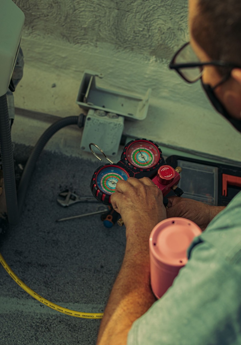 Close-up of hands holding a refrigerant manifold gauge set beside an air conditioning unit.