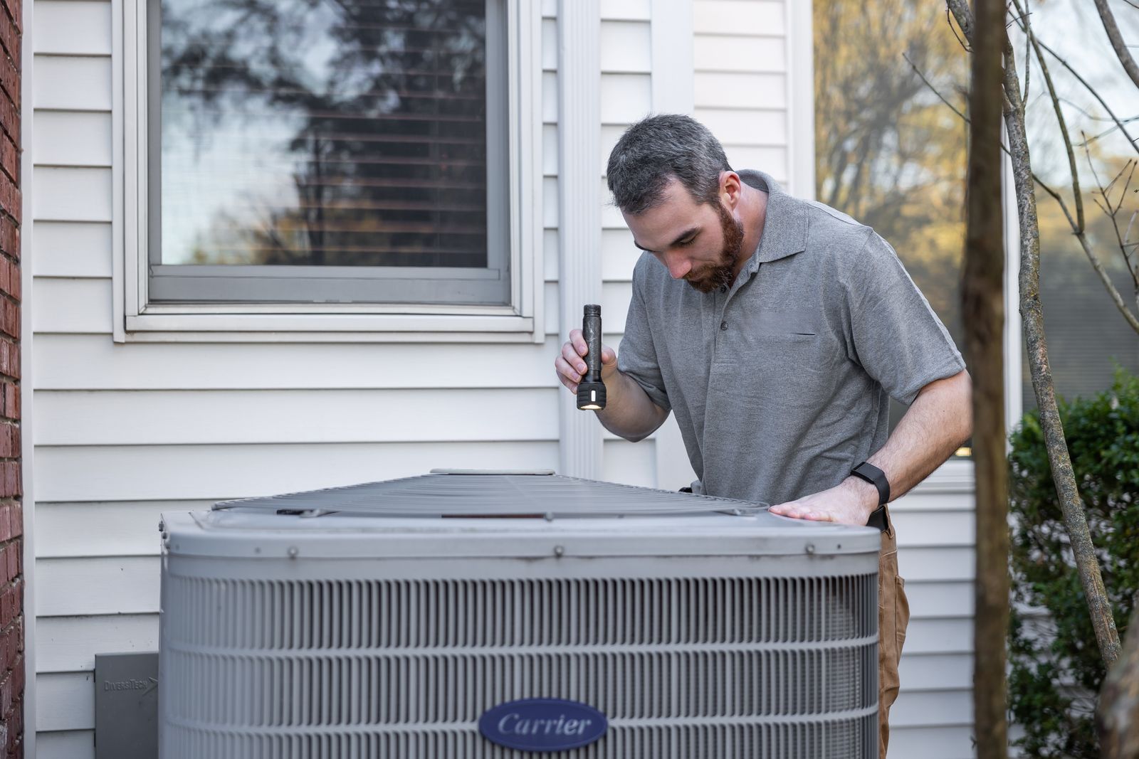 A technician with a flashlight inspecting the top of an outdoor air conditioning condenser beside a house.